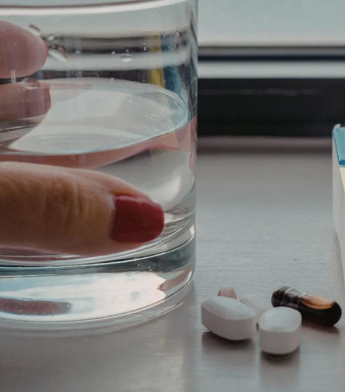 Hand holding a glass of water beside a pill organizer and scattered pills on a table