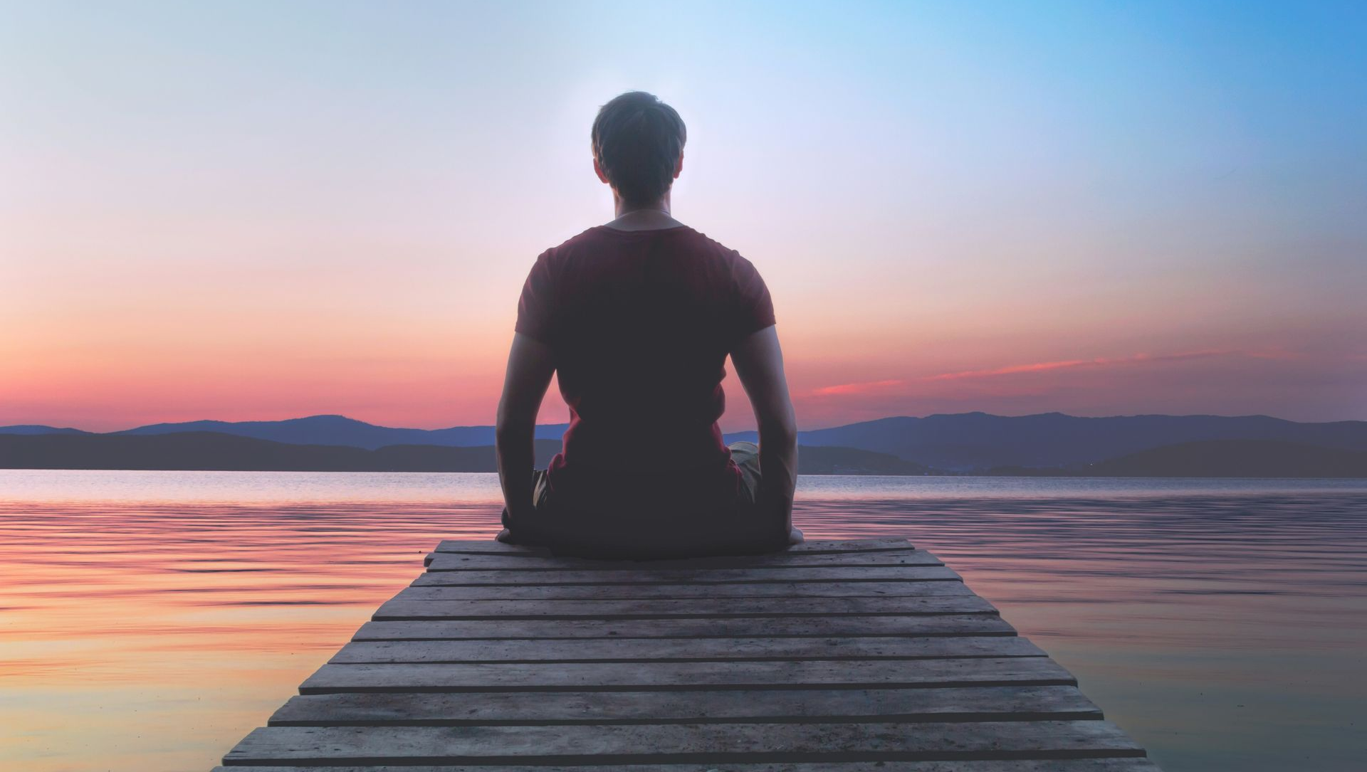Person sits on a wooden dock at sunset, facing a calm lake under a pink-blue sky.