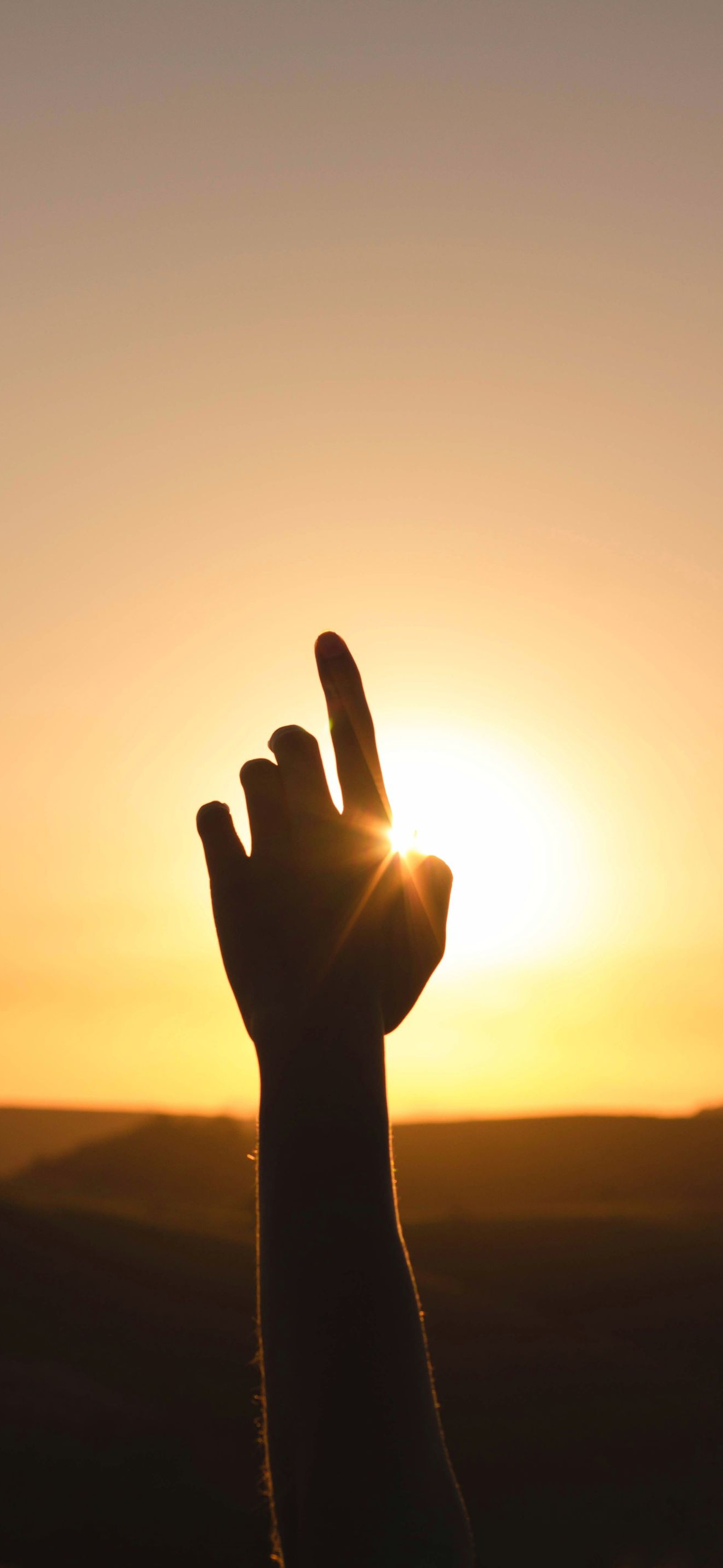 Silhouette of a raised hand reaching toward the sun at sunset over a dark landscape