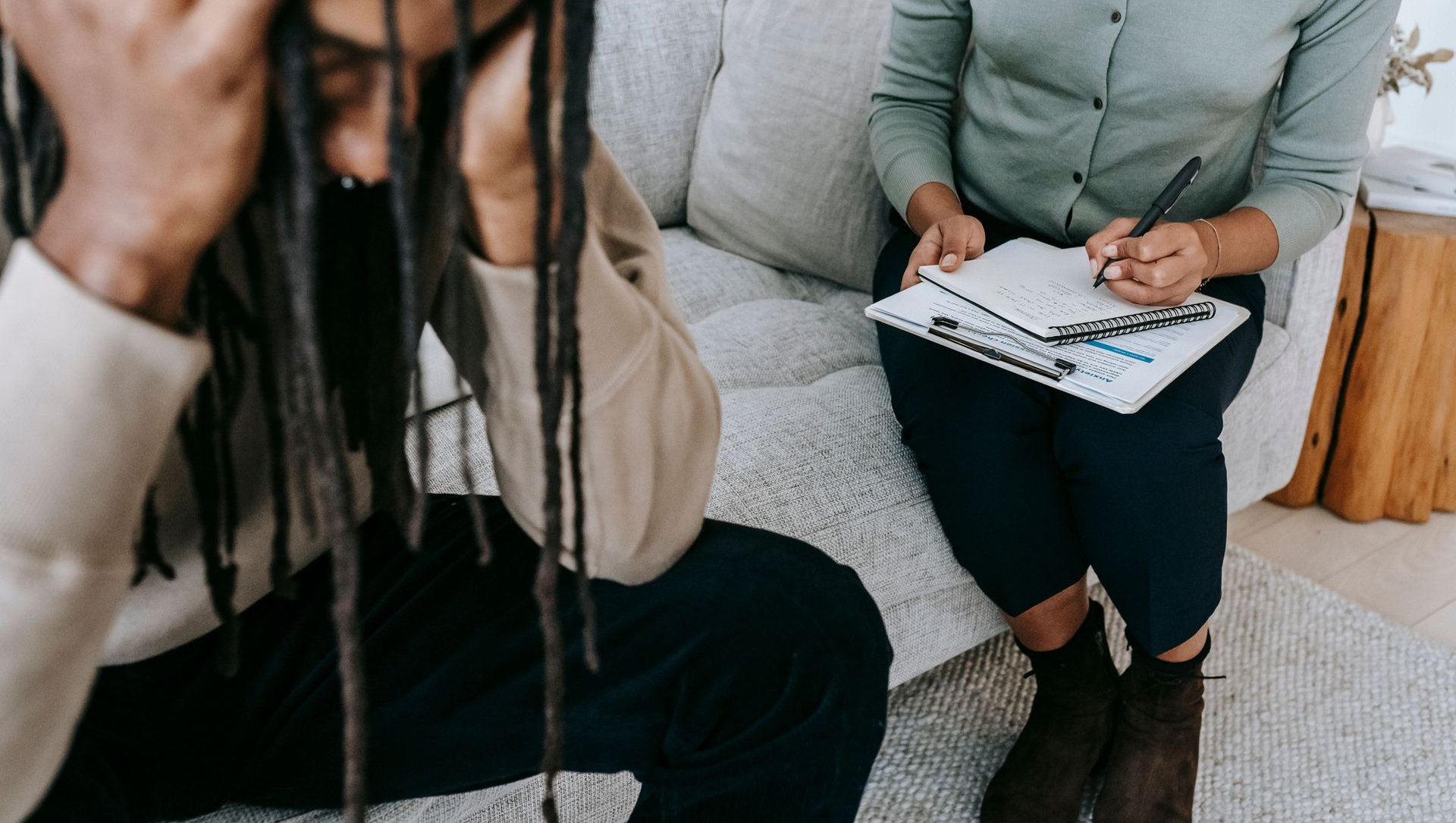 Two people talking on a couch, one holding a clipboard and pen.