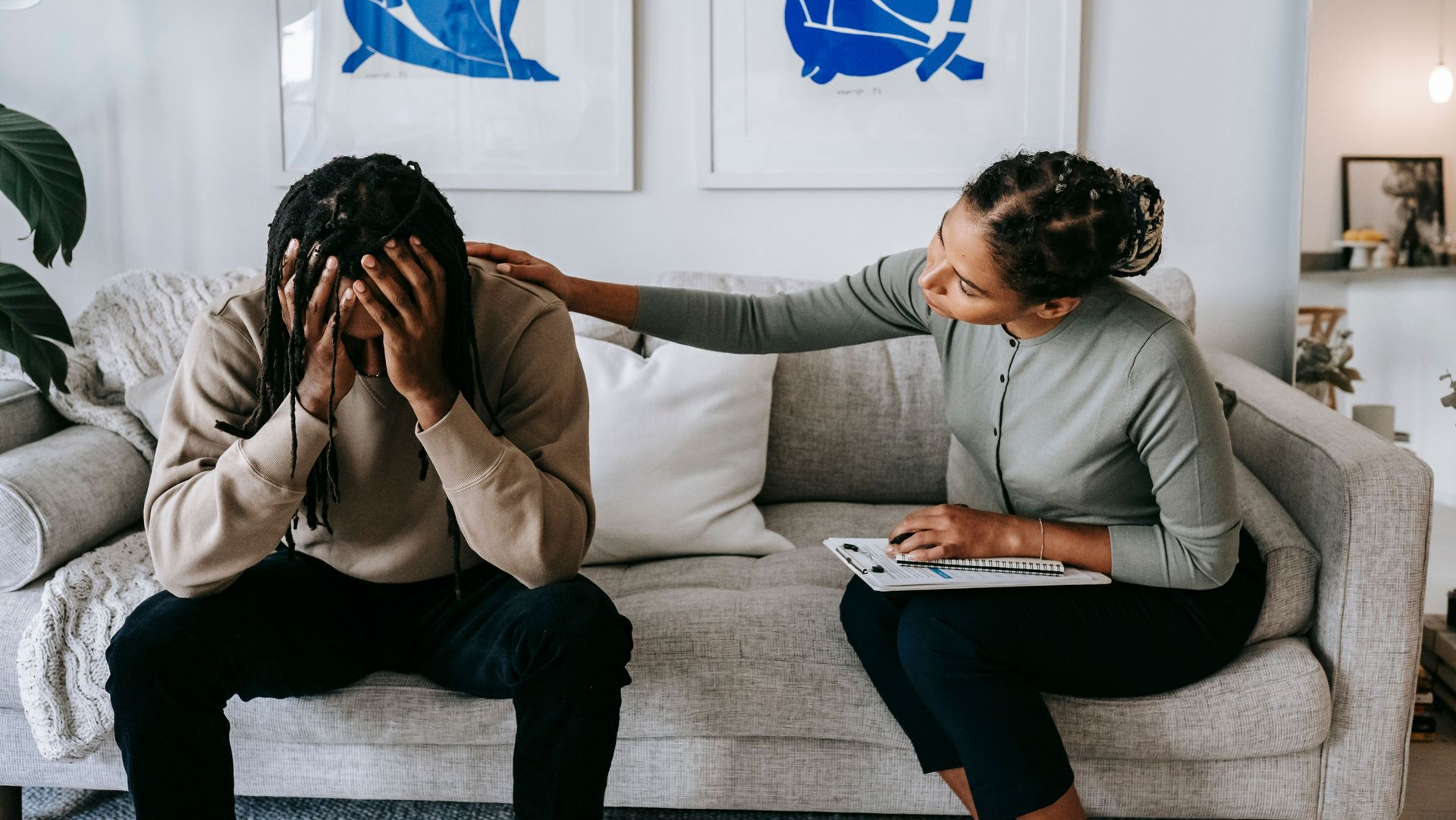Person sits on couch with head in hands as another comforts them in a bright living room.