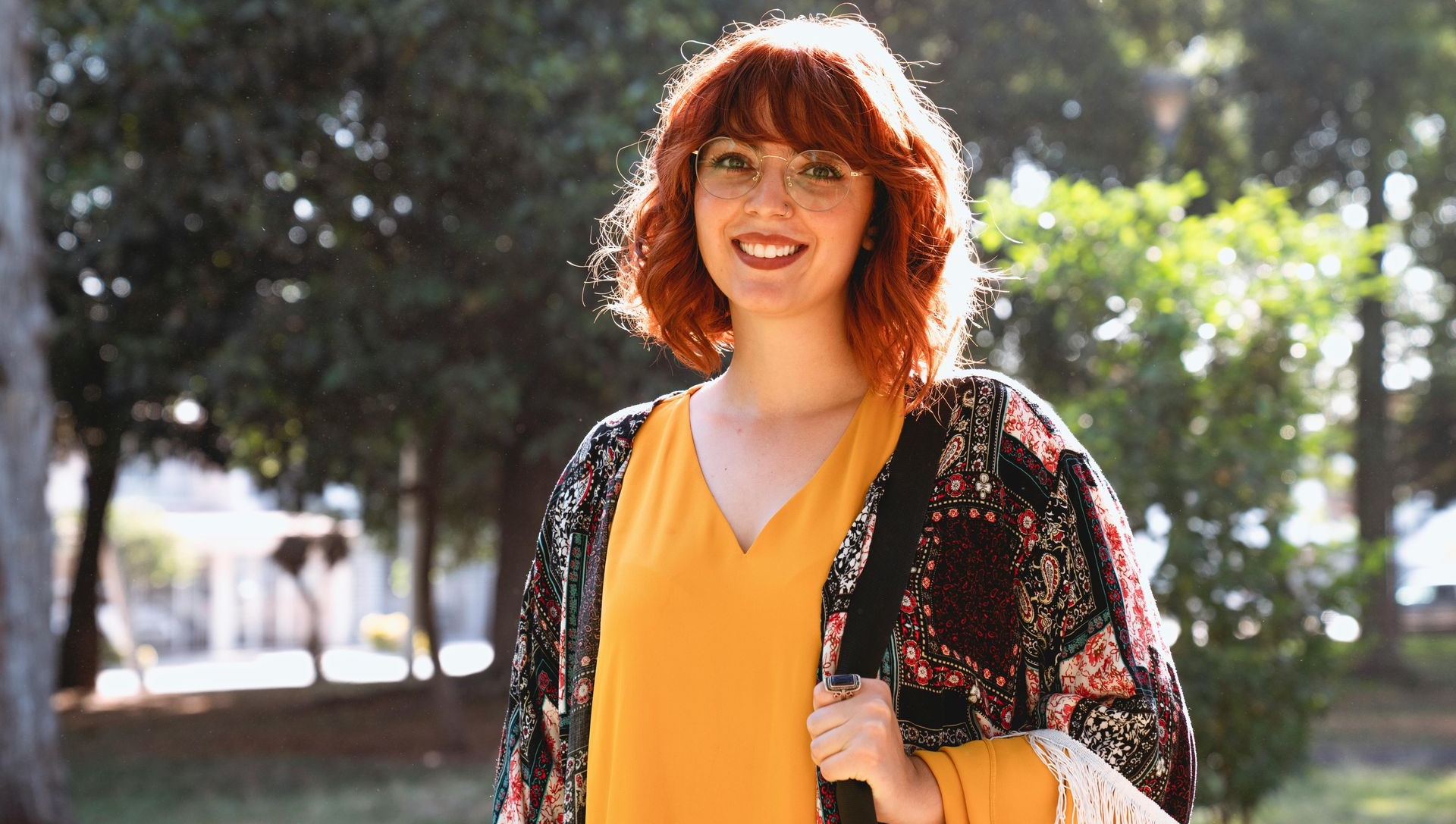 Smiling woman with red hair in a yellow dress and floral cardigan standing outdoors in sunlight
