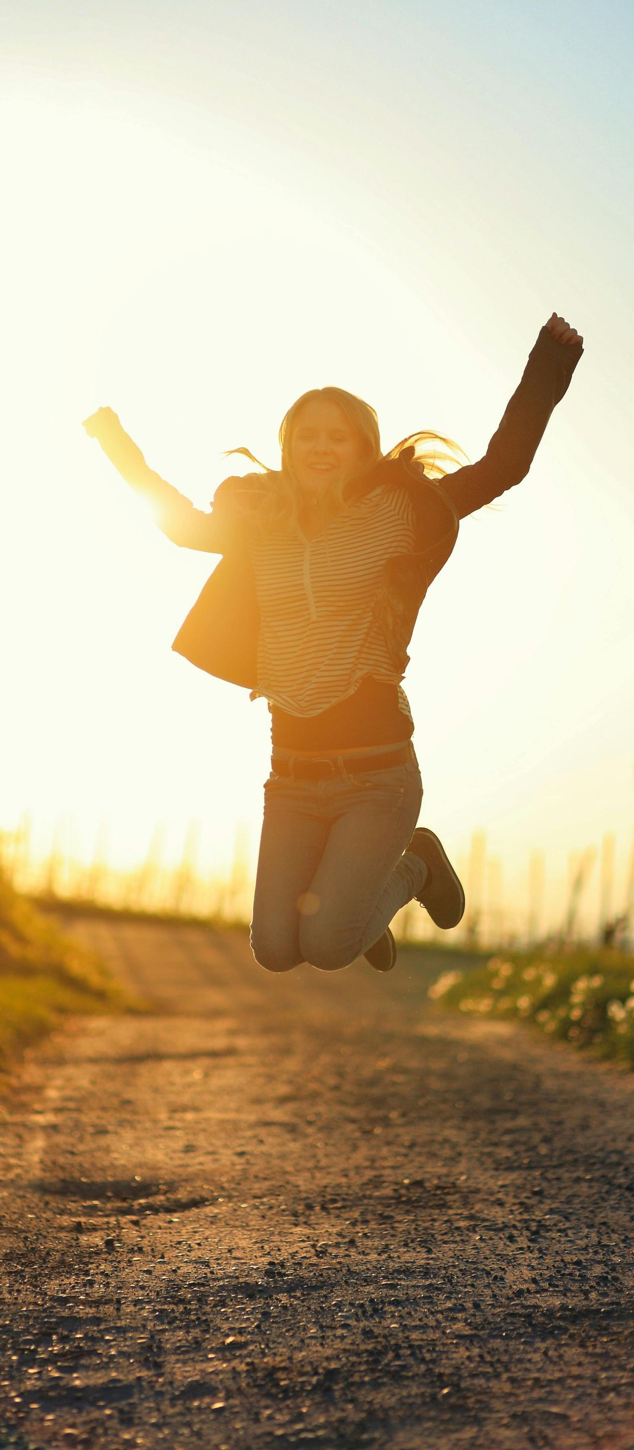 Person jumping on a dirt path at sunset, arms raised, backlit by golden light.