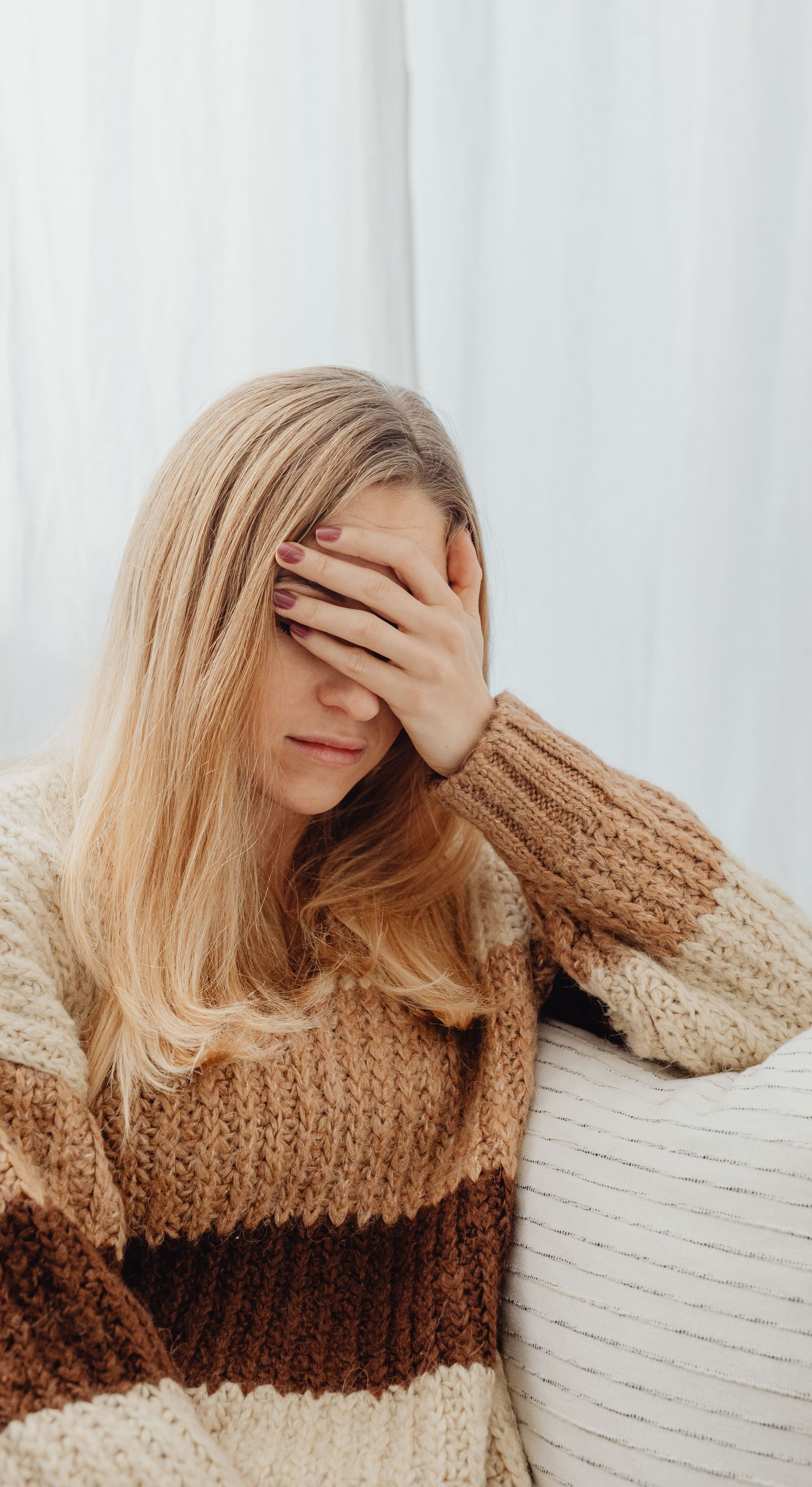 Blonde person in a tan sweater covers their face while sitting on a couch by a white curtain.