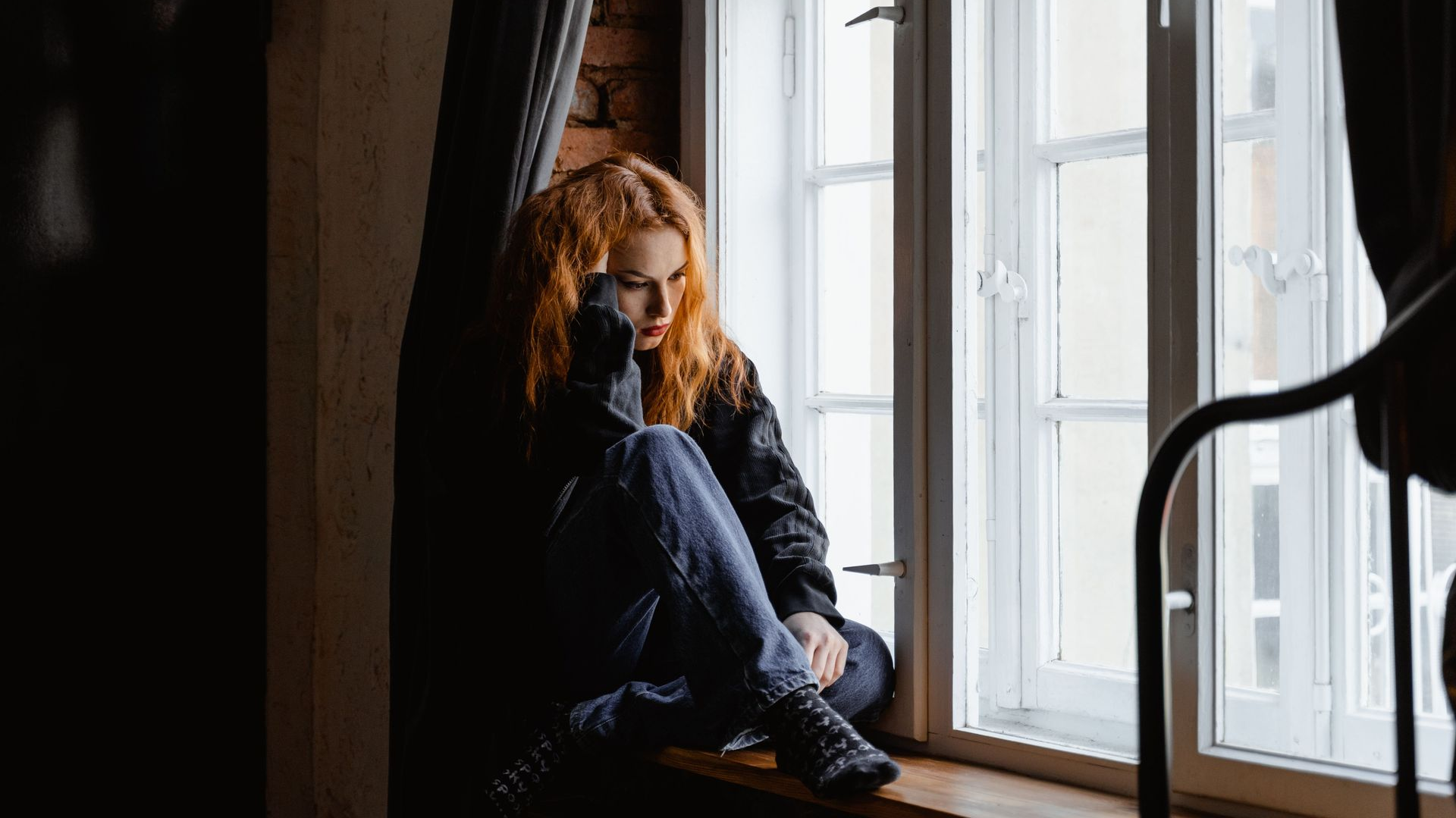 Person sitting by a bright window, looking contemplative in a dark, cozy room.