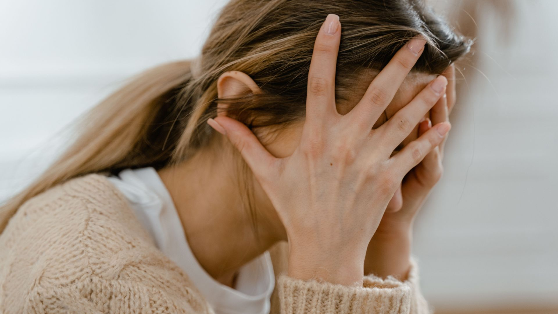 Distressed person covering their face with both hands while sitting indoors in a beige sweater
