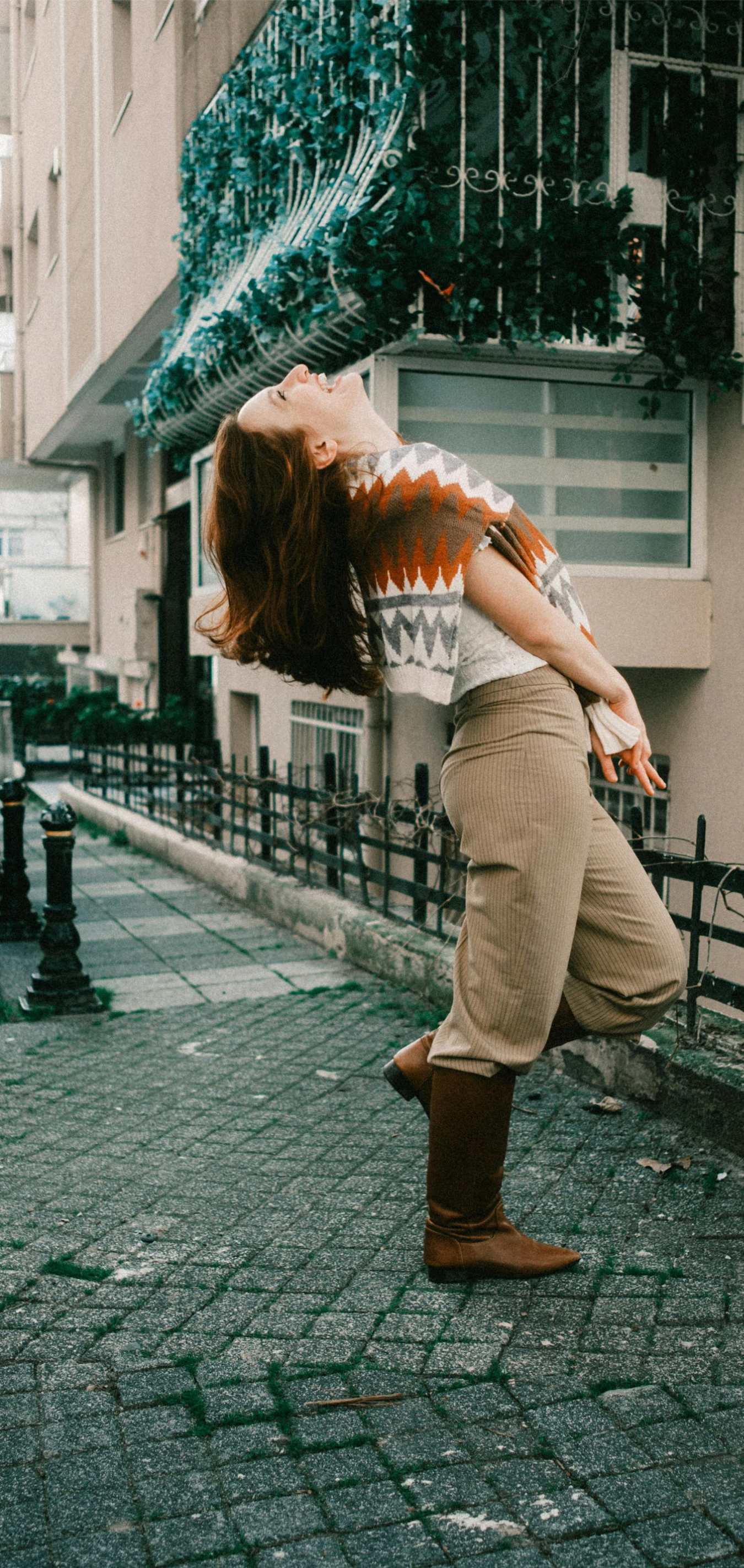 Person leaning back on a wet cobblestone street beside a building, wearing beige clothes and brown boots.