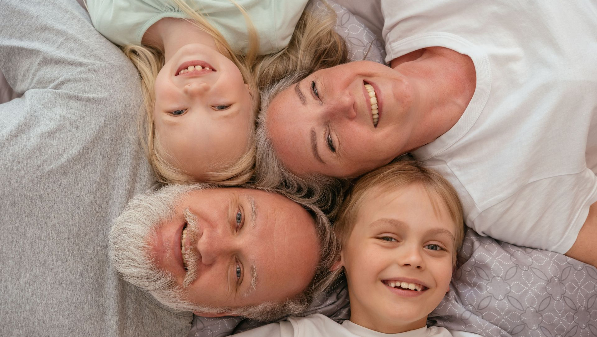 Four smiling people lying head-to-head, looking up at the camera on a soft gray surface