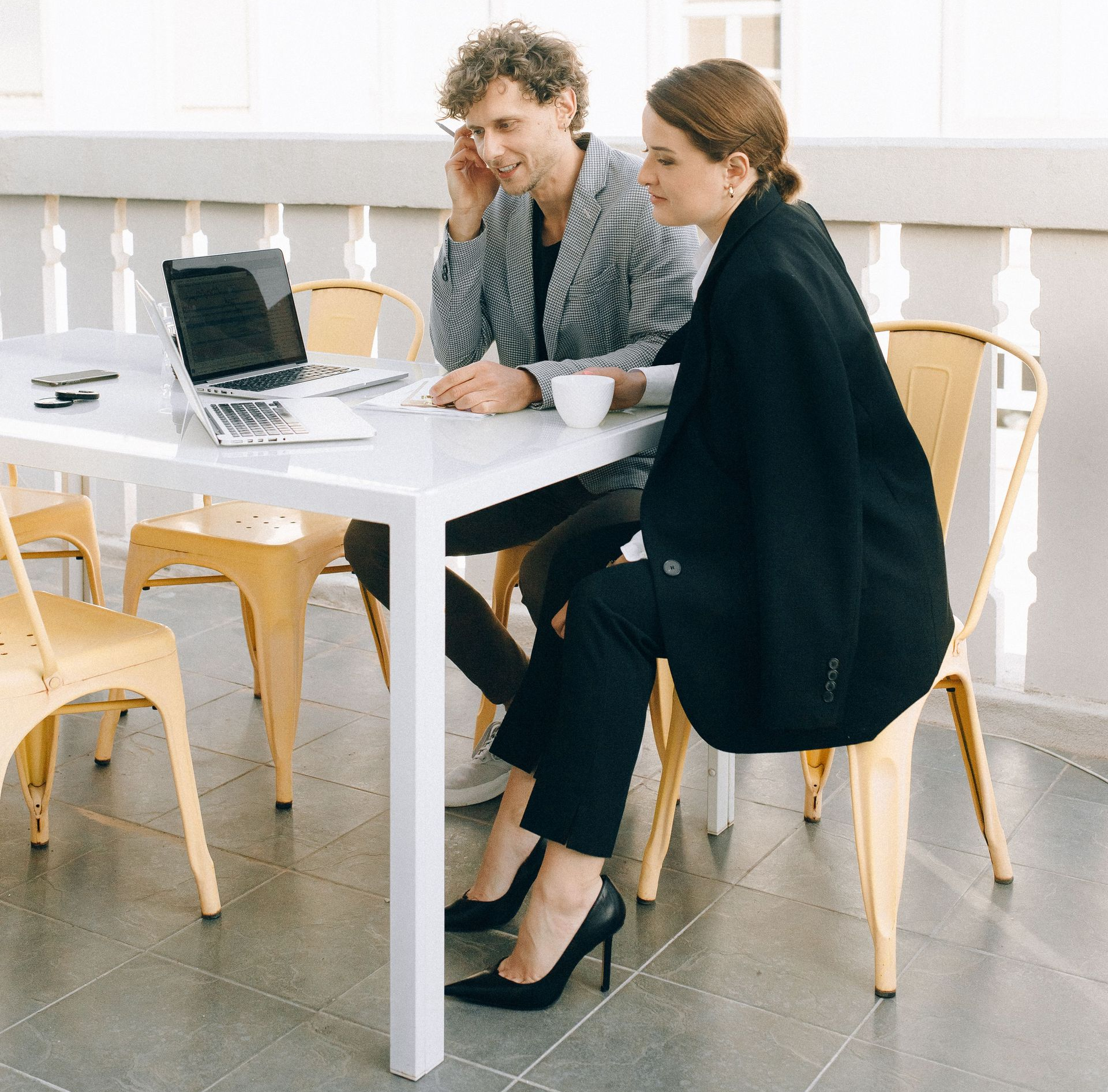 Two people working at a white outdoor table with a laptop in a bright courtyard