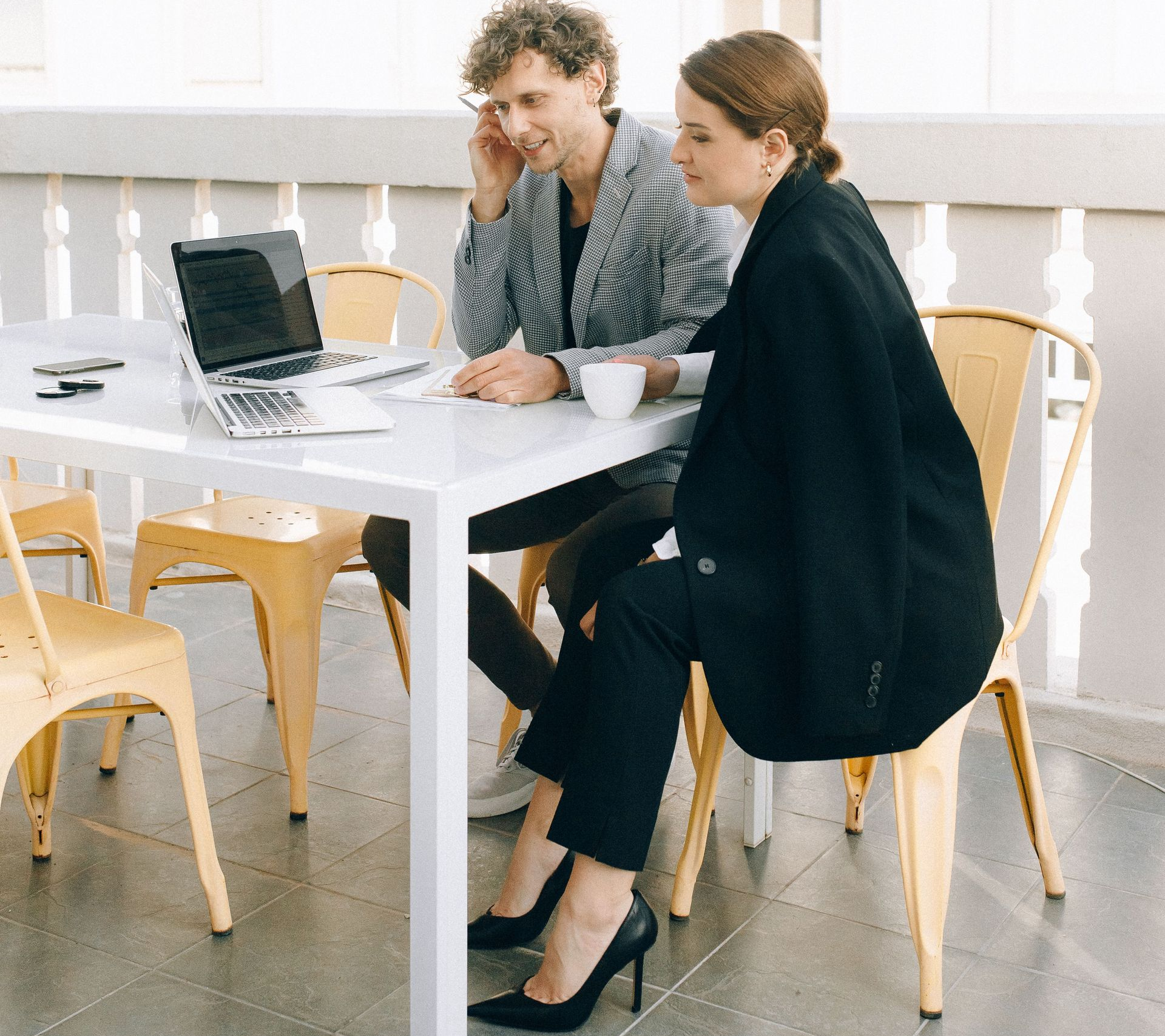 Two coworkers in a bright office discussing work at a white table with a laptop open.