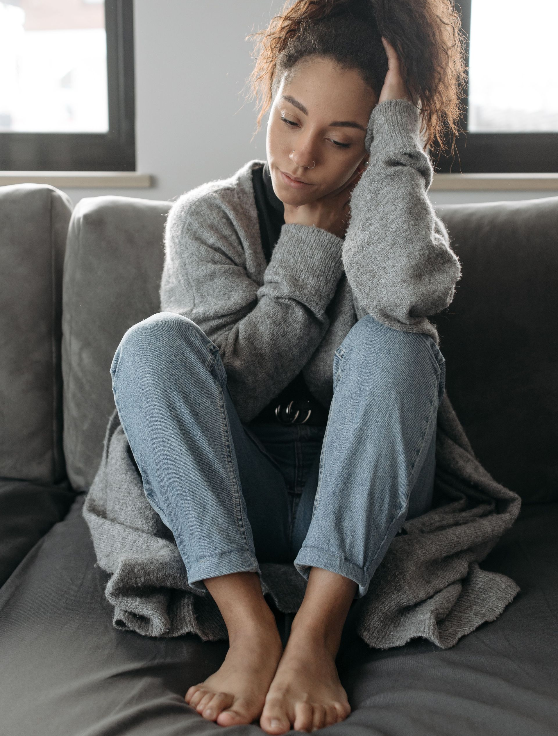 Person sitting on a couch, knees hugged to chest, looking down thoughtfully in a gray sweater and pants.