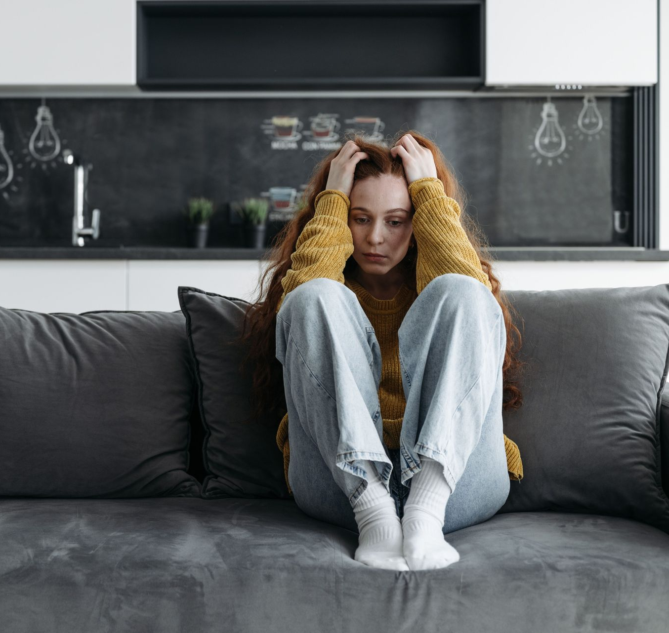 Person sitting curled up on a gray sofa in a modern kitchen, looking down, with a somber expression.