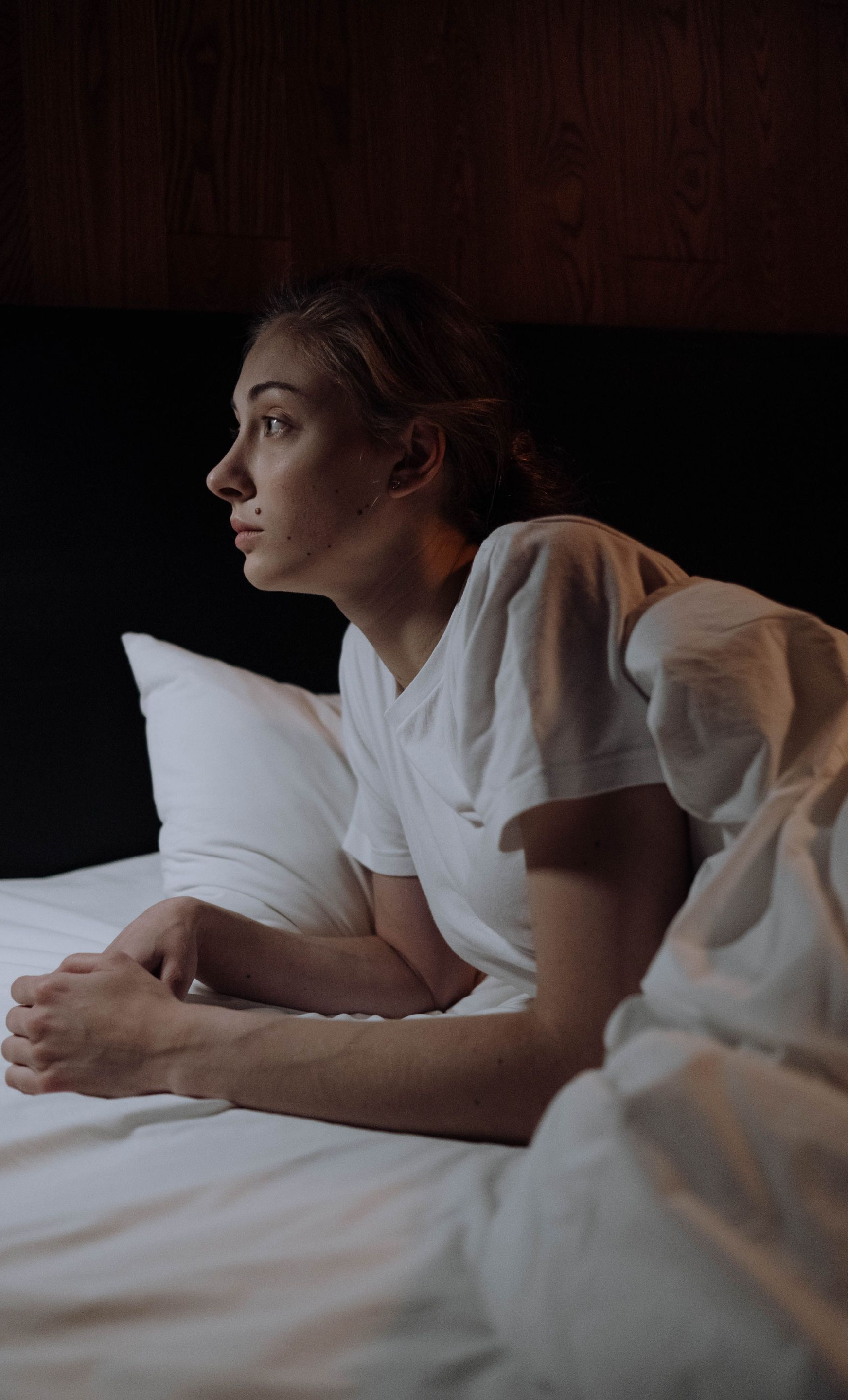 Person in a white shirt lying on a bed, resting on a pillow in dim light