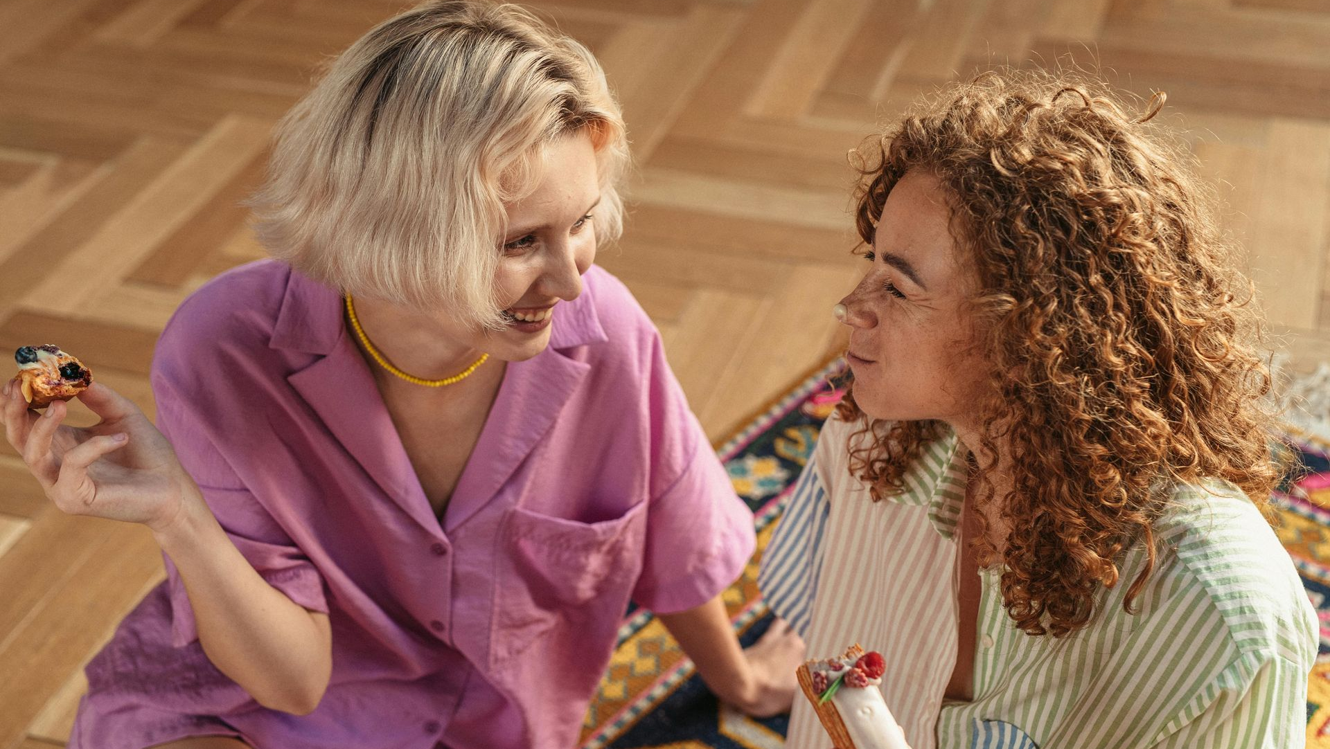 Two people sitting on a wooden floor, talking and holding food, one in pink and one in green stripes.