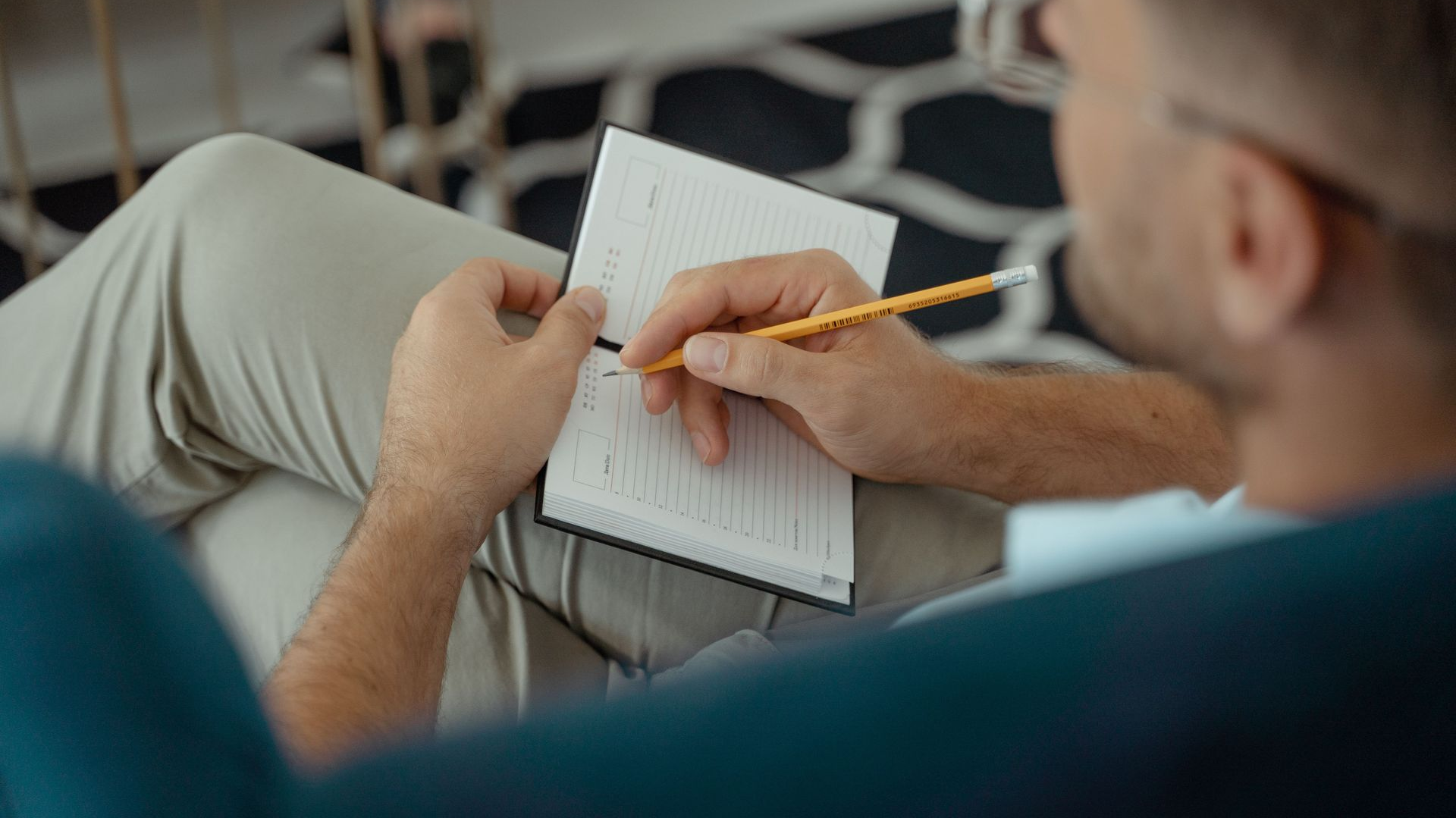 Person writing notes in a notebook on a couch, holding a yellow pencil.