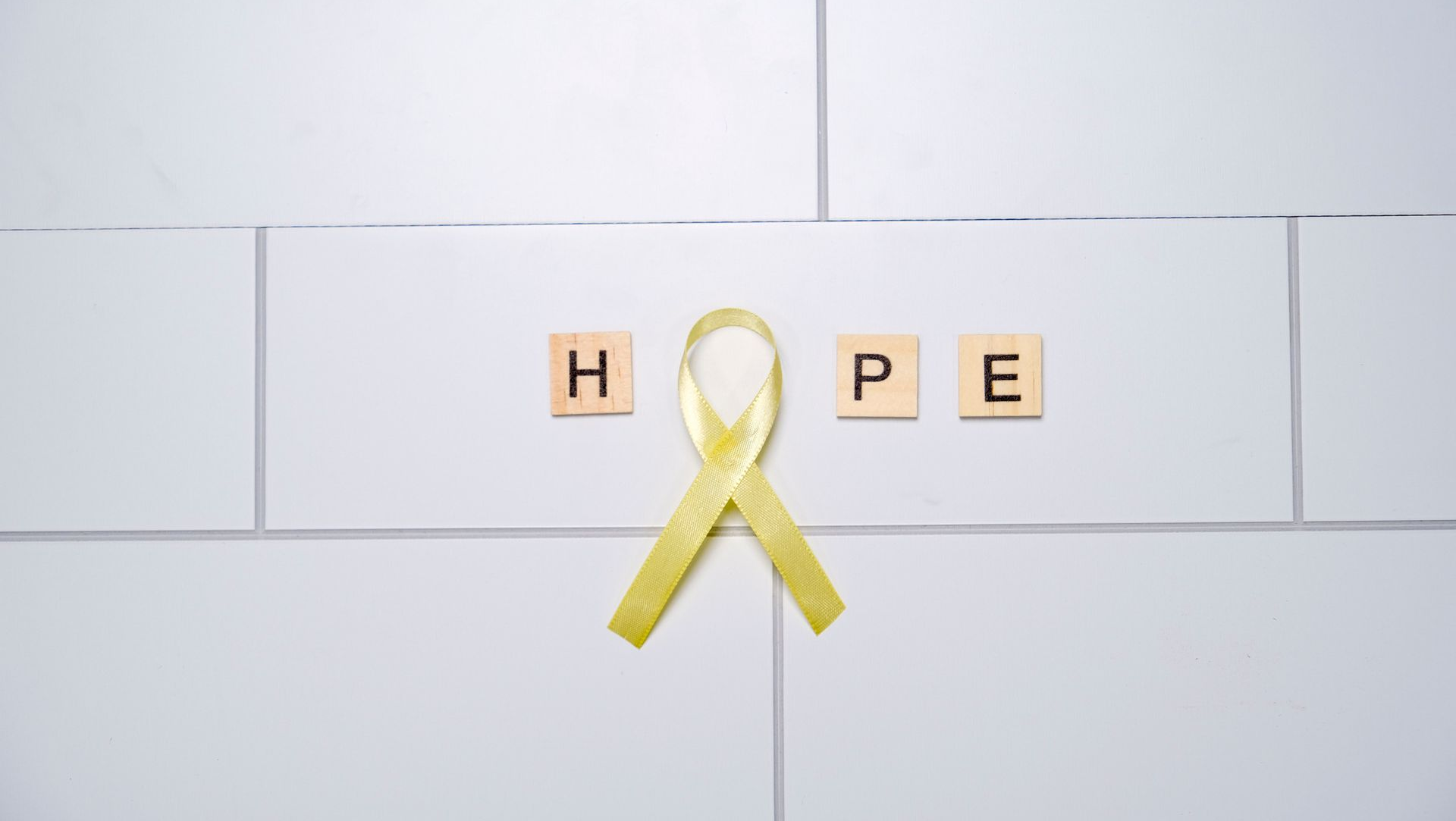 Yellow ribbon over wooden letters spelling “HOPE” on a white tiled wall.