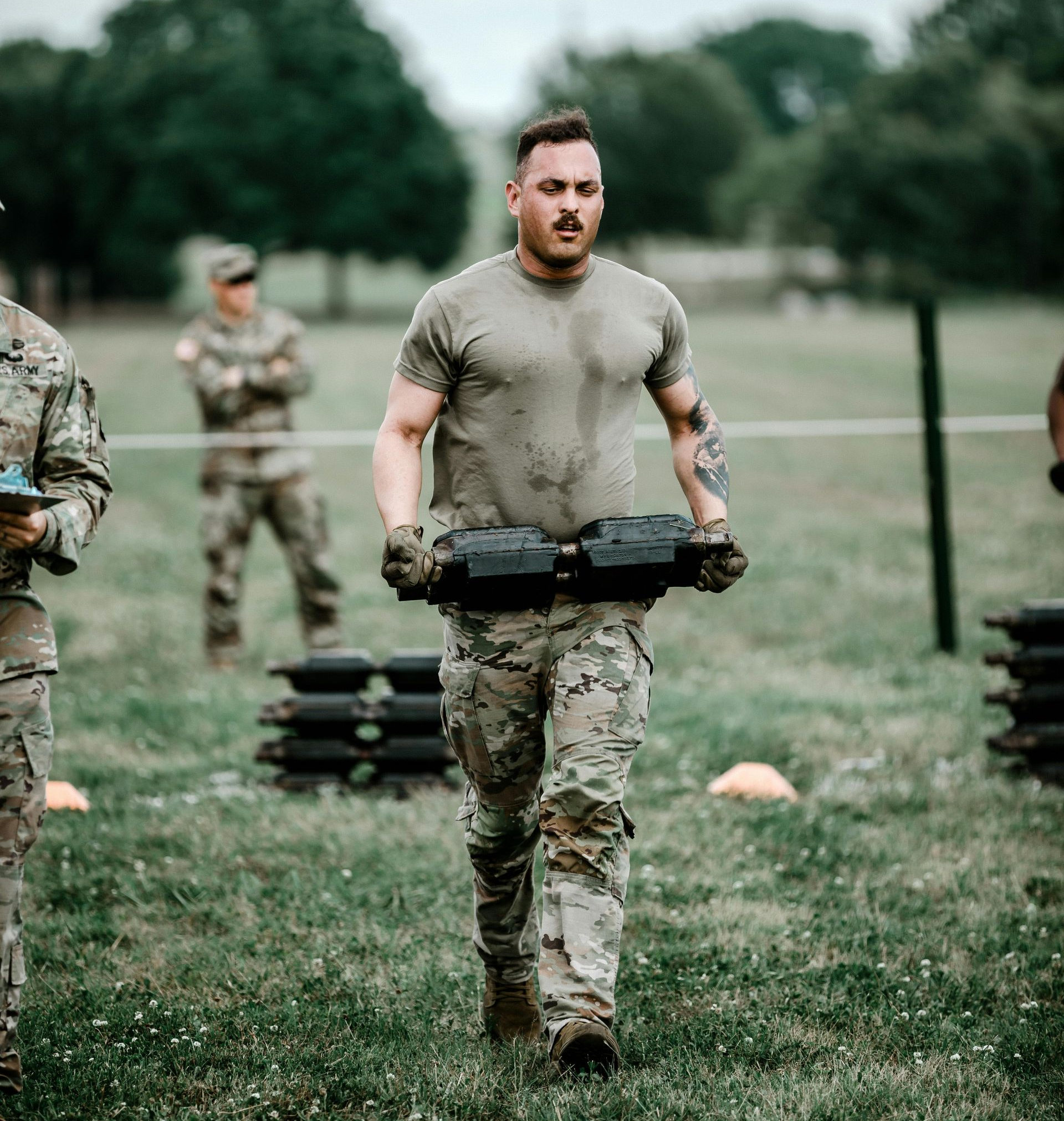 Soldier in camo carrying weights outdoors, other soldiers and obstacle course in background.