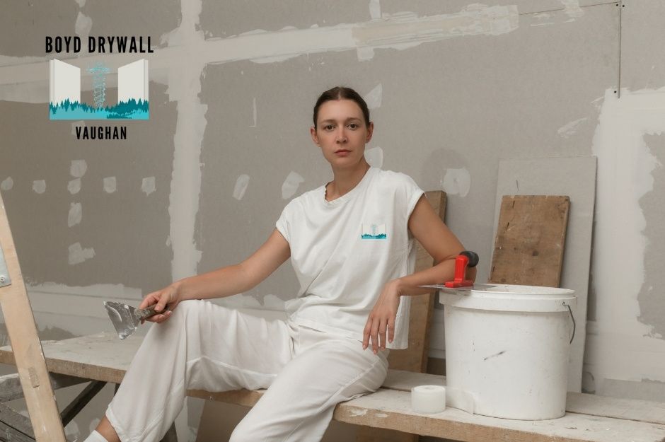 A drywall repair company professional in a white outfit sitting on a wooden bench, holding a putty knife, surrounded by unfinished drywall walls with joint compound patches, paint buckets, and construction materials in a commercial workspace near Durham Regional Rd 12, and Trans - Canada Hwy, Brock, ON.
