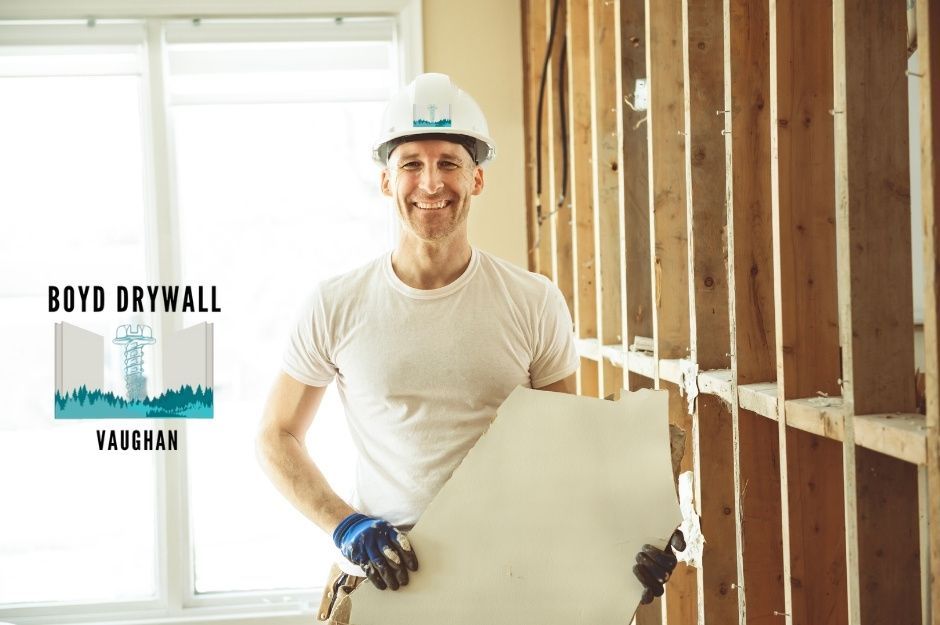 A drywall repair company worker wearing a white hard hat and a t-shirt, smiling while holding a drywall sheet at a commercial construction site with exposed wooden wall studs and bright window lighting near Delrex Blvd, and Guelph St, Halton Hills, ON.