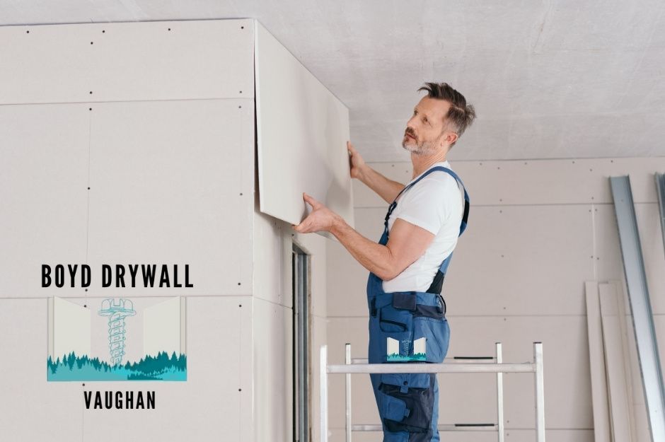 A drywall repair company professional in blue overalls installs a white drywall panel on an interior wall, carefully positioning it with both hands while standing on a work platform in a commercial space near Livingston St, and Wellington St, Pickering, ON.
