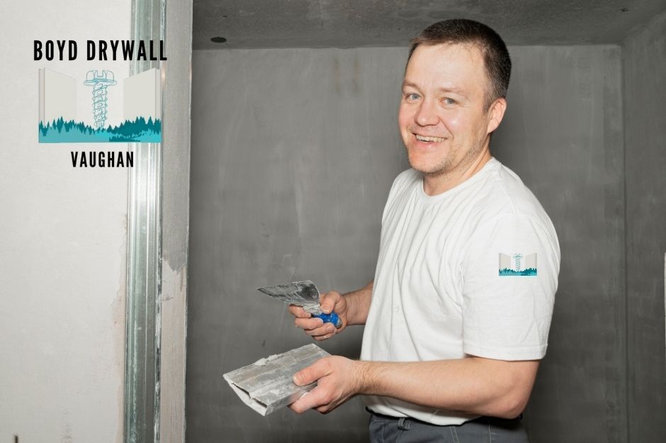 A drywall repair contractor in a white shirt smiles while applying joint compound to a gray drywall surface using a putty knife in a residential construction project, working in an unfinished room near Woodstream Ave, and Grandview St N, Oshawa, ON.