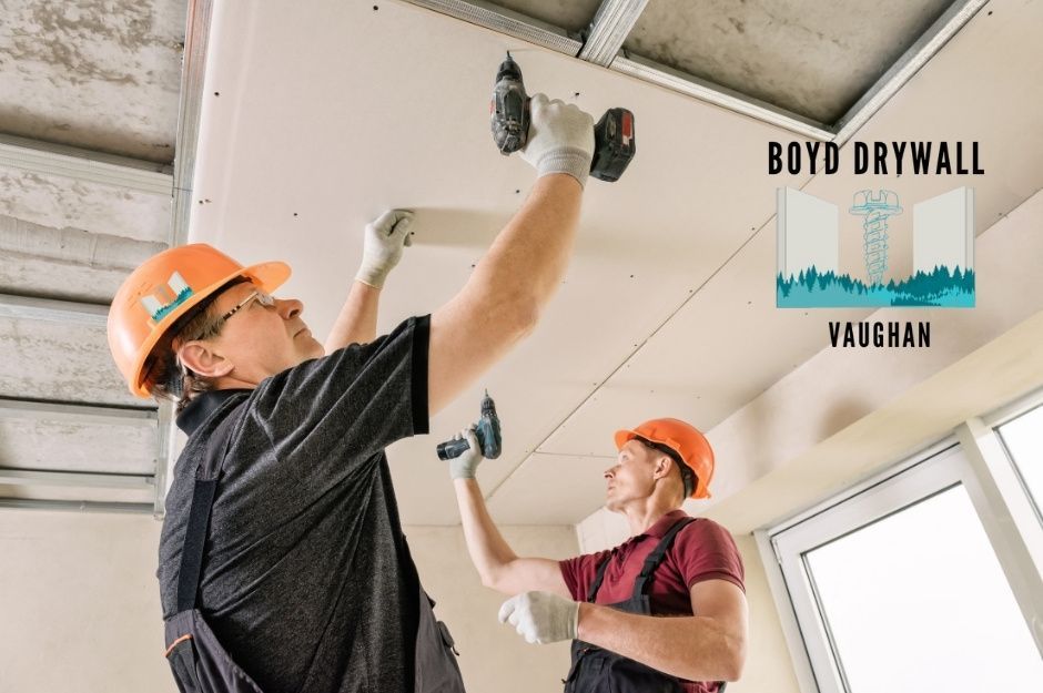 Drywall repair specialists wearing orange hard hats and work gloves install drywall ceiling panels using cordless drills at a residential construction site. Two workers collaborate on the overhead installation near Chapman Dr, and Coles Ave, Ajax, ON.