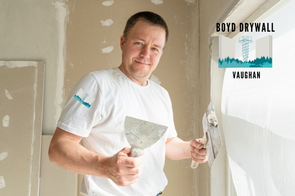 A drywall repair technician wearing a white t-shirt smiles while holding two putty knives at a residential construction site with unfinished drywall walls showing joint compound patches. The professional is working on wall finishing near Whitby Shores Greenway, and Point Hope Pl, Whitby, ON.