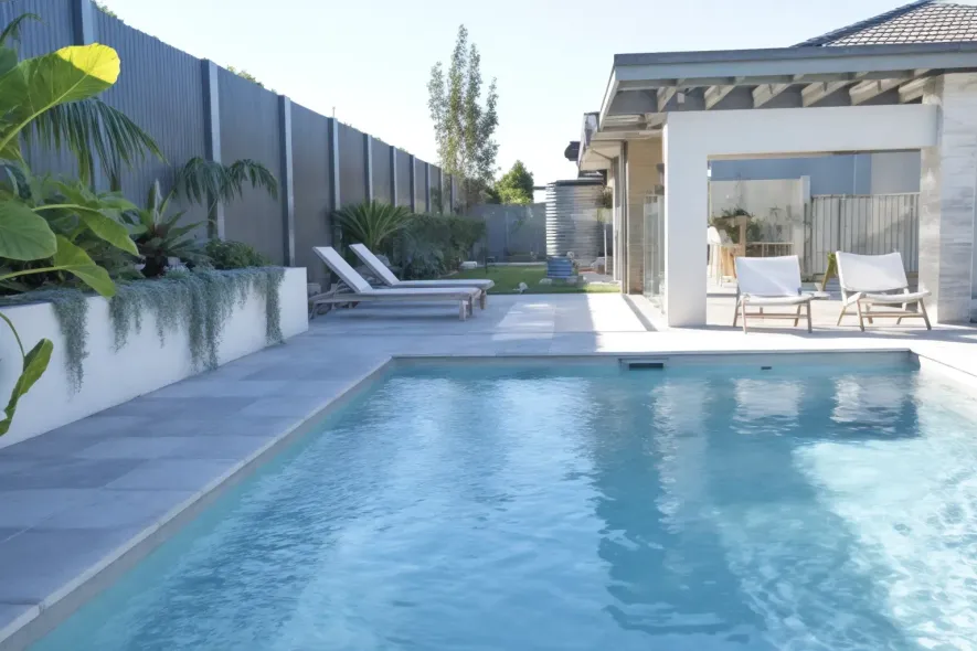 A rectangular pool with light blue water, surrounded by gray paving, lounge chairs, and a modern house.