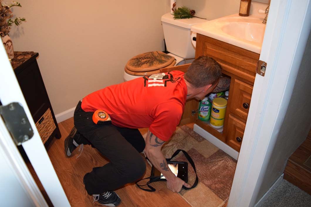 A man in a red shirt is kneeling down in a bathroom looking under a sink.