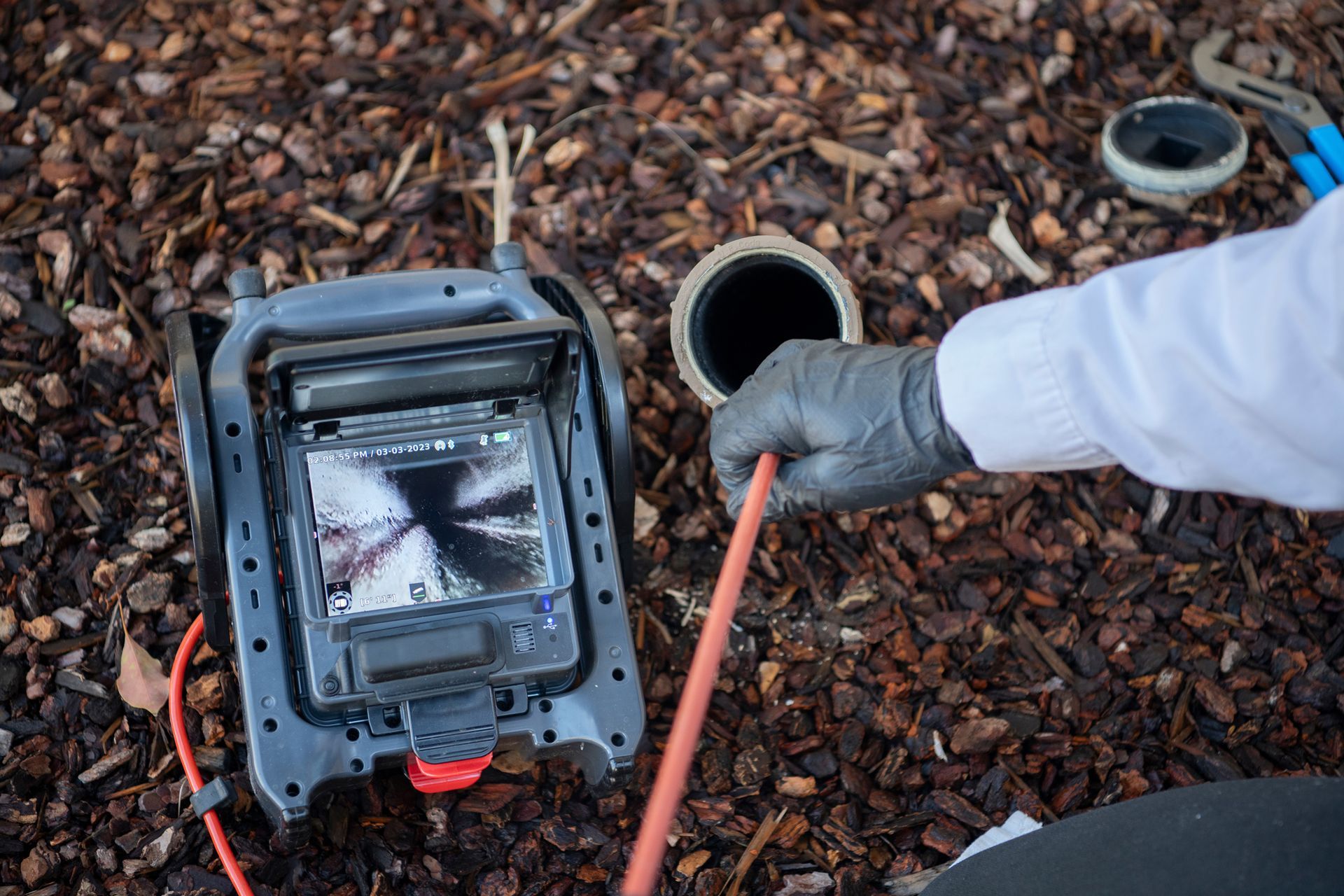 Worker in gloves using a drainage inspection camera beside open pipes on wood chips