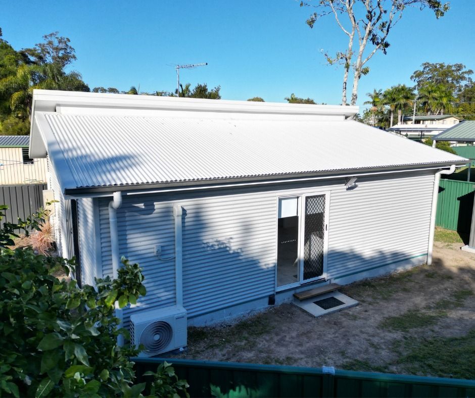 Gray corrugated-iron-clad building with a white roof and black framed doors, set in a yard.