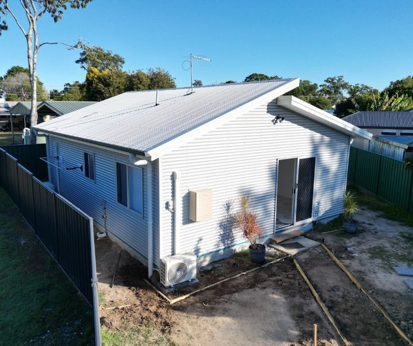 A small, light-colored house with a metal roof and a fenced yard under a blue sky.