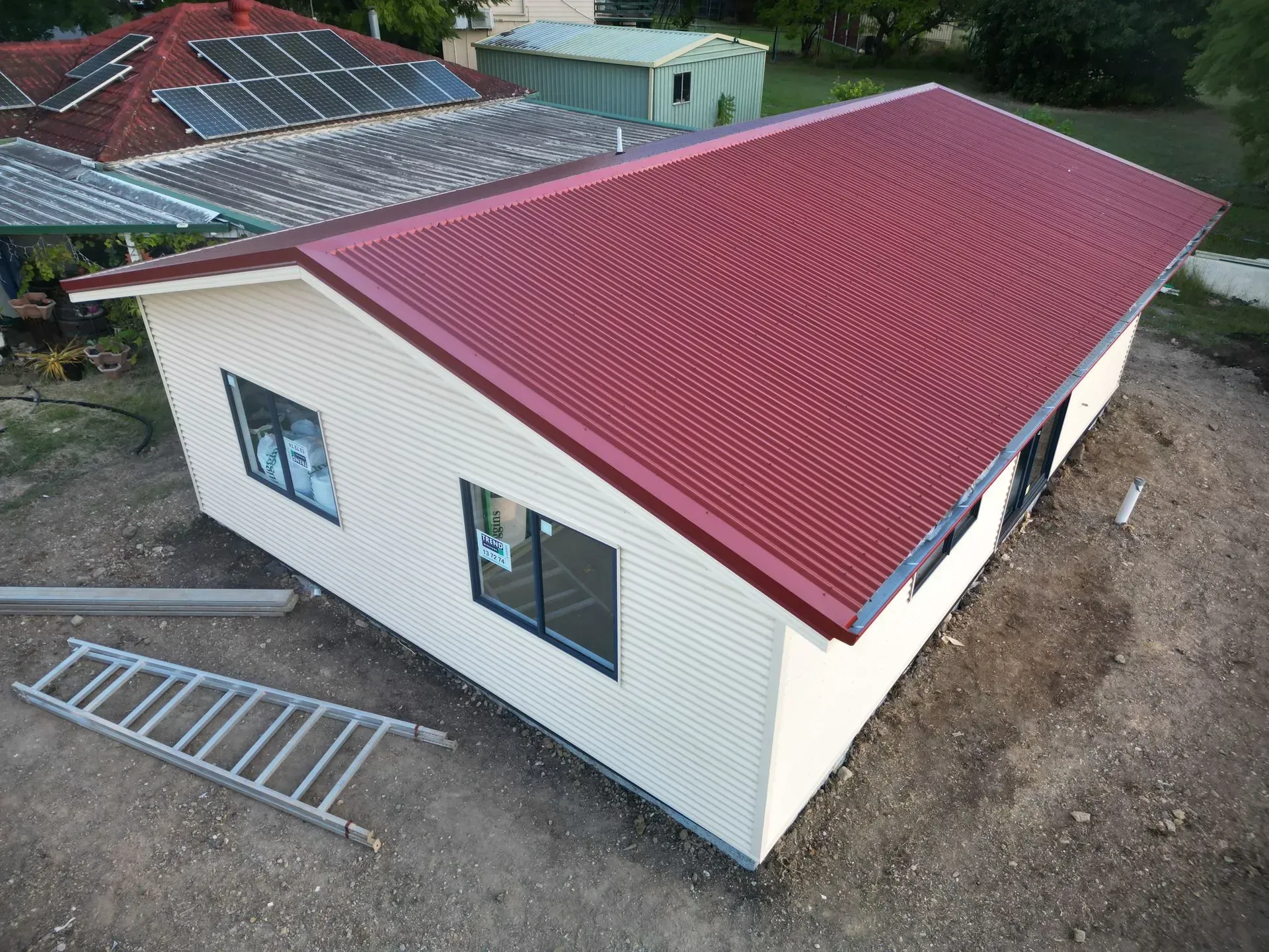 Small building with red corrugated metal roof, cream siding, windows, and ladder on the ground.