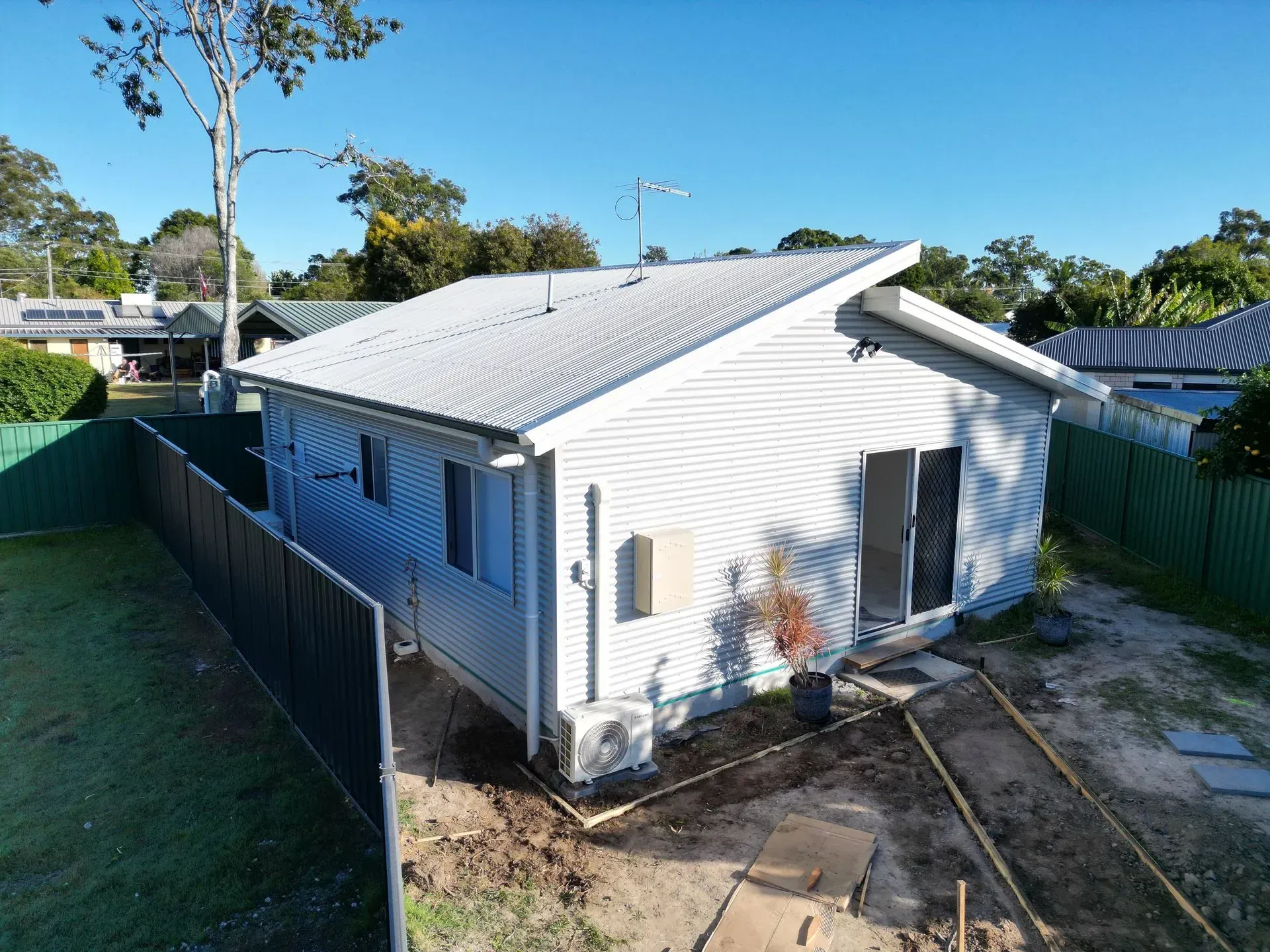 White house with angled roof and side entrance; surrounded by fence and trees on a sunny day.