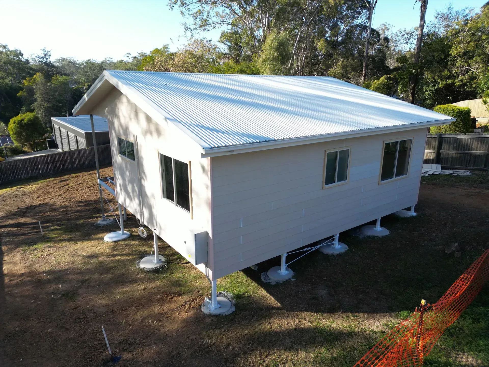 Beige house elevated on concrete supports, corrugated metal roof, windows, and surrounding brown yard.