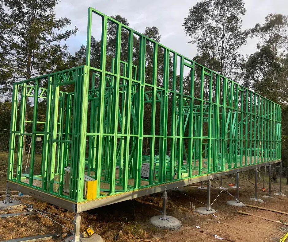 Green wood framing for a house under construction, supported by metal posts on concrete footings.