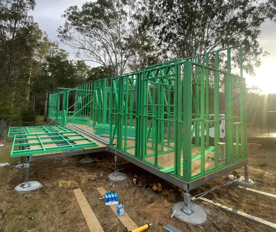 Green metal-framed house under construction on concrete piers. Timber floor is visible. Outdoors, daytime.