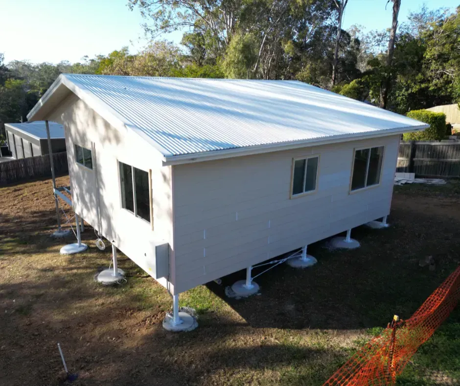 Elevated light-colored building with a corrugated metal roof, sitting on concrete piers in a grassy area with trees in the background.