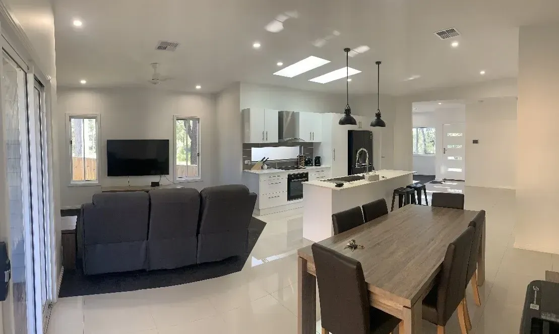 Open-plan living space with a kitchen, dining area, and living room. Neutral color palette with gray sofa and wood table.