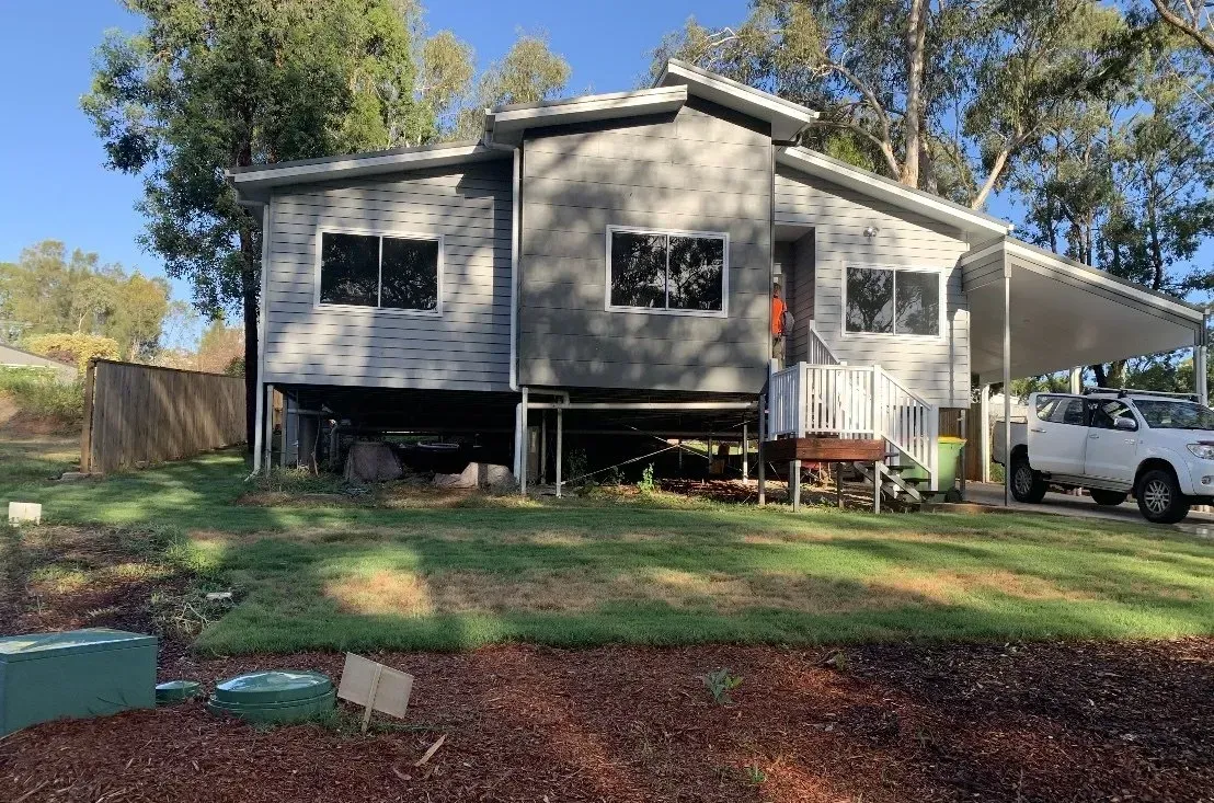 Elevated gray house with windows and carport, on a grassy lot, parked white truck.
