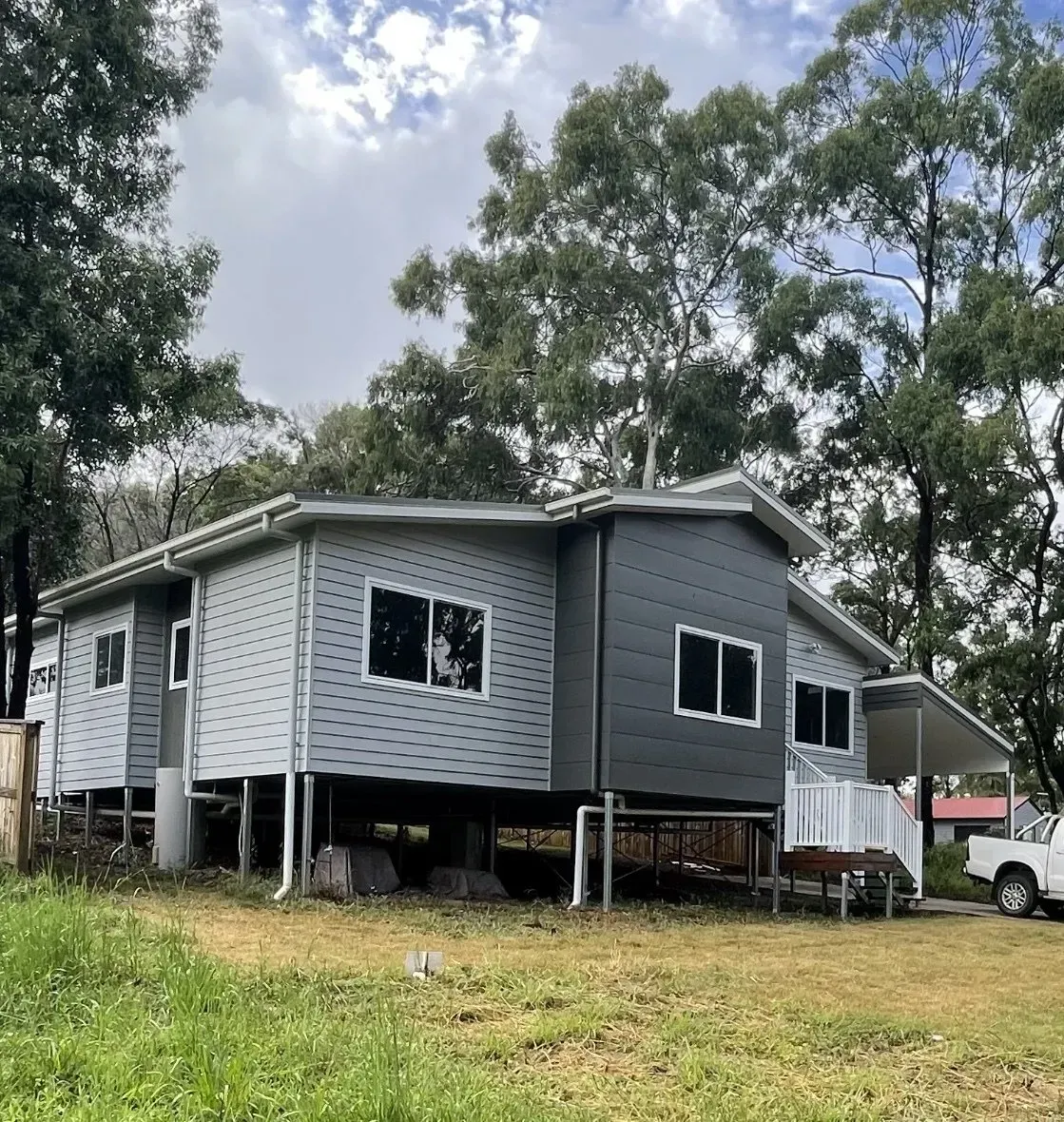 A light gray house with dark gray trim and white support beams, set in a grassy area with trees.