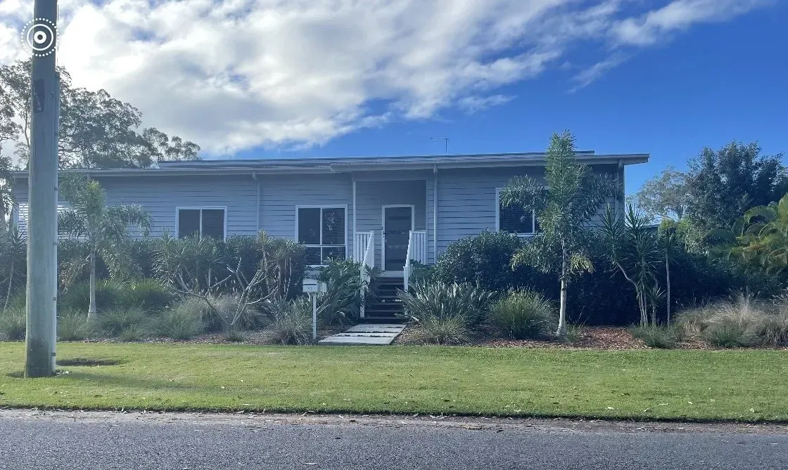 Gray house with porch, steps, and windows, surrounded by trees and grass, under a cloudy sky.