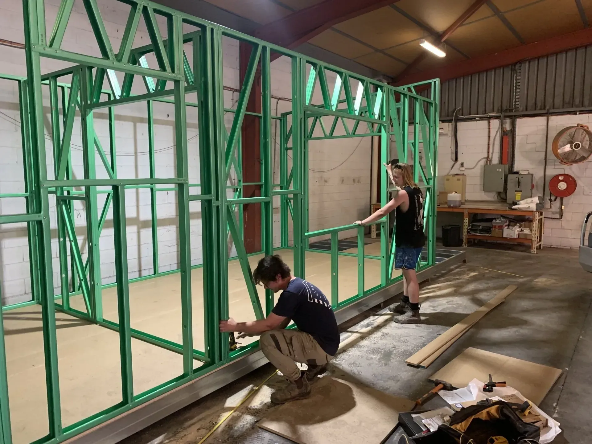 Two people building a green framed wall inside a workshop. One is kneeling.