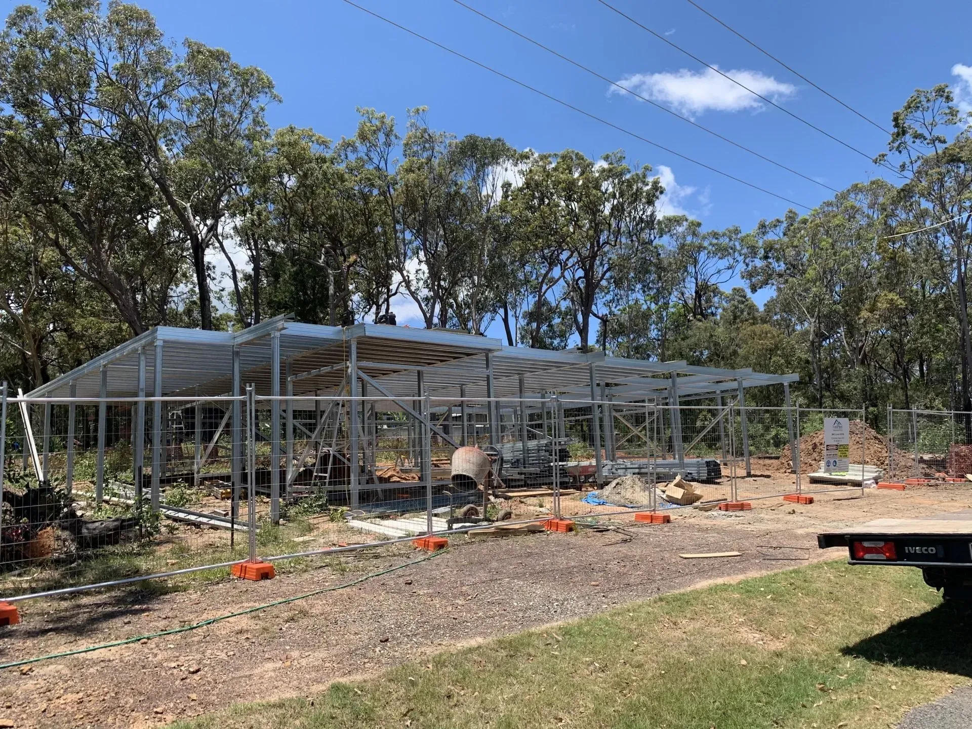Metal frame of a building under construction, outdoors with trees and a cloudy sky.