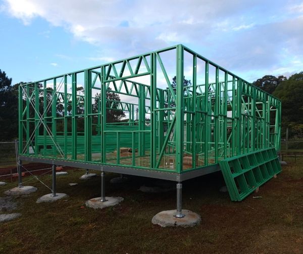 Green-framed house under construction on concrete piers, set in a grassy outdoor area under a cloudy sky.