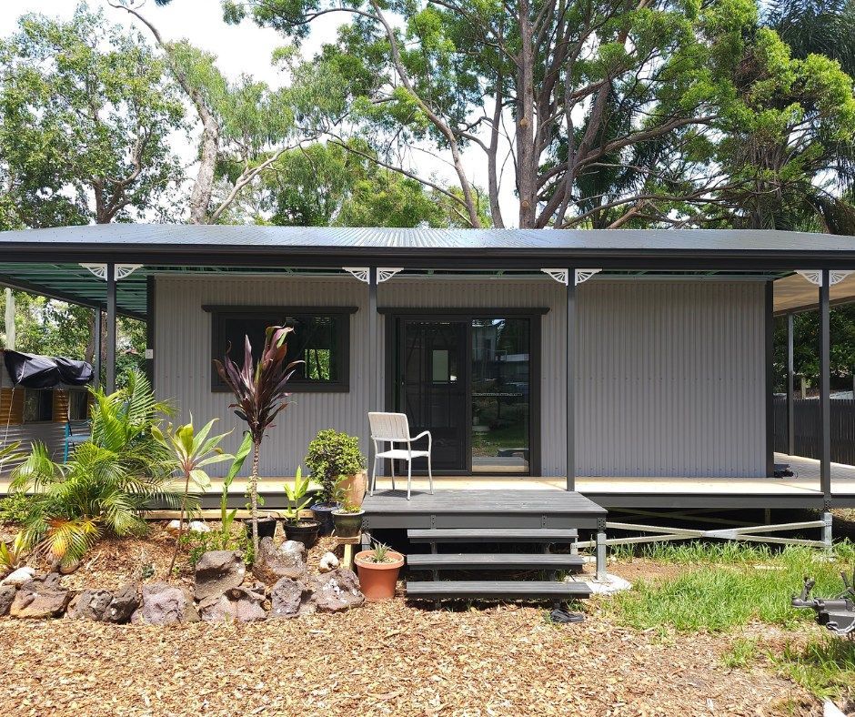 Gray modular home with dark trim, porch, and canopy, surrounded by trees and landscaping.
