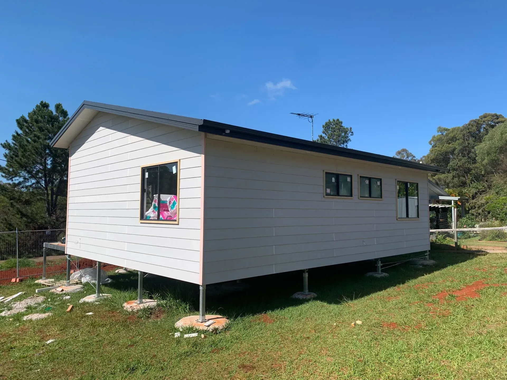 Small, light-colored house elevated on stilts with a dark roof. Green grass and blue sky surround.