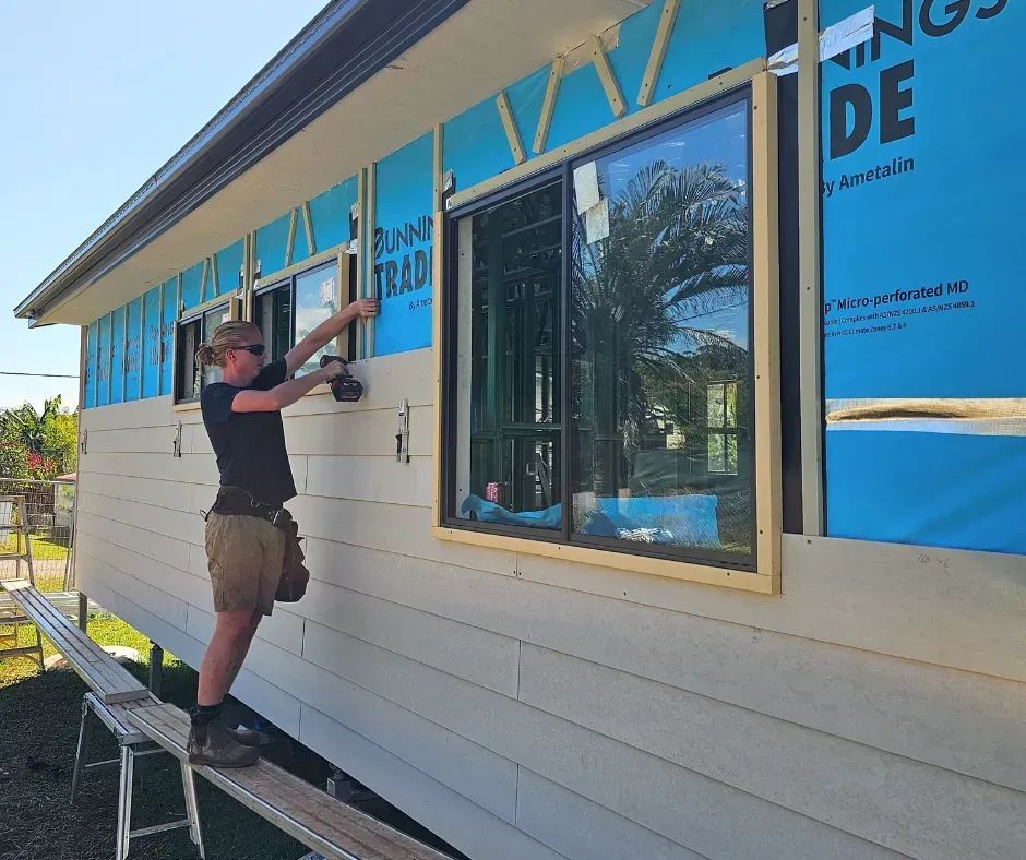 Man installing siding on a house, standing on a scaffold next to a window.