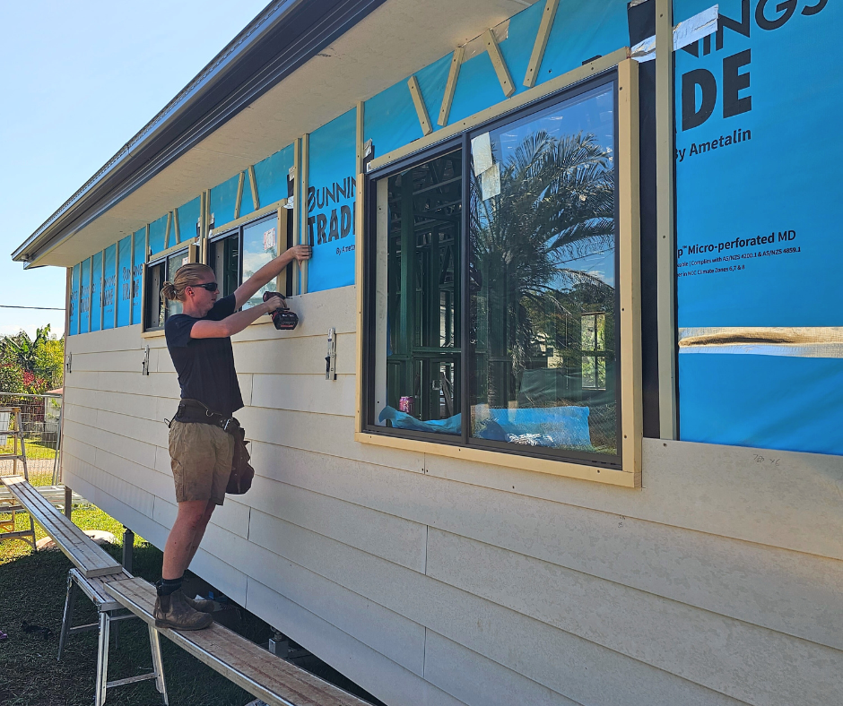 A person installing siding on a house, standing on a step. Blue wrap around windows. Sunny outdoors.