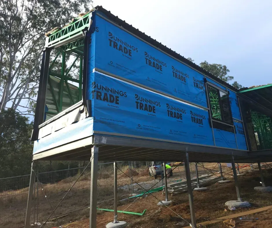 House under construction on stilts, wrapped in blue weatherproofing.
